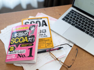 窓際の木製デスクの上に適正検査SCOAの参考書2冊、PCとメガネ、ペンが置いてあるところ